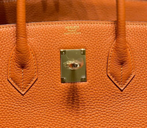 HERMÈS Birkin. Exquisite Craftsmanship and Timeless Elegance: Discover the Allure of the HERMES Birkin Bag Large Leather Top Handle. Designed with the actress Jane Birkin in mind, the Birkin bag is a contemporary development of the Kelly bag. Created based on Jane's suggestions, it was architected to be more significant to fit modern women's fast-paced life, with several minor yet outstanding adaptations, such as the elongated top handles. The French Birkin 30 bag is a classic touch of elegance to your look! The accessory features a trapeze body, a pebbled leather texture, a hanging key fob, gold-tone hardware, round top handles, a padlock fastening detail, a foldover top with twist-lock closure, a front center logo stamp, an internal zipped pocket, and purse feet. | CRIS AND COCO Authentic Quality Luxury Accessories