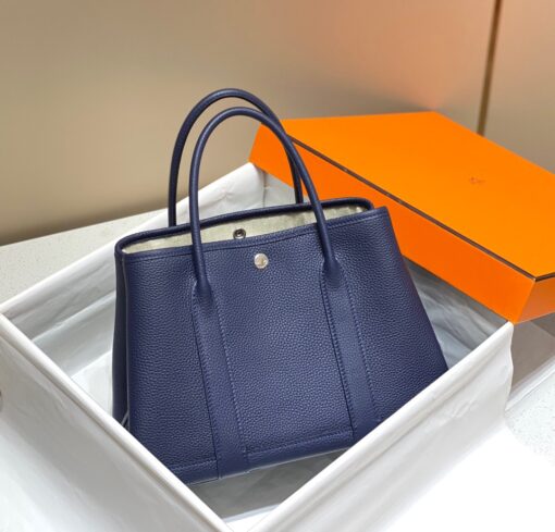 HERMÈS Garden Party: Style for Every Occasion. This Garden Party tote is the perfect blend of style and practicality. This chic and sporty bag is designed for every occasion, whether spending the day amongst the hustle and bustle of the city or enjoying a peaceful weekend in the countryside. Crafted with both leather and canvas, it's the perfect choice for whatever your destination. Not only is it spacious enough to hold your everyday essentials, but it also features Clou de Selle snap closures on the sides for added convenience. This snap that allows the bag to be just the right amount of malleable; to expand and create extra interior space to accommodate larger, or more items at once. Wherever your adventures take you, this exclusive HERMÈS tote will accompany you every step of the way. | CRIS&COCO Authentic Quality Designer Bags and Luxury Accessories